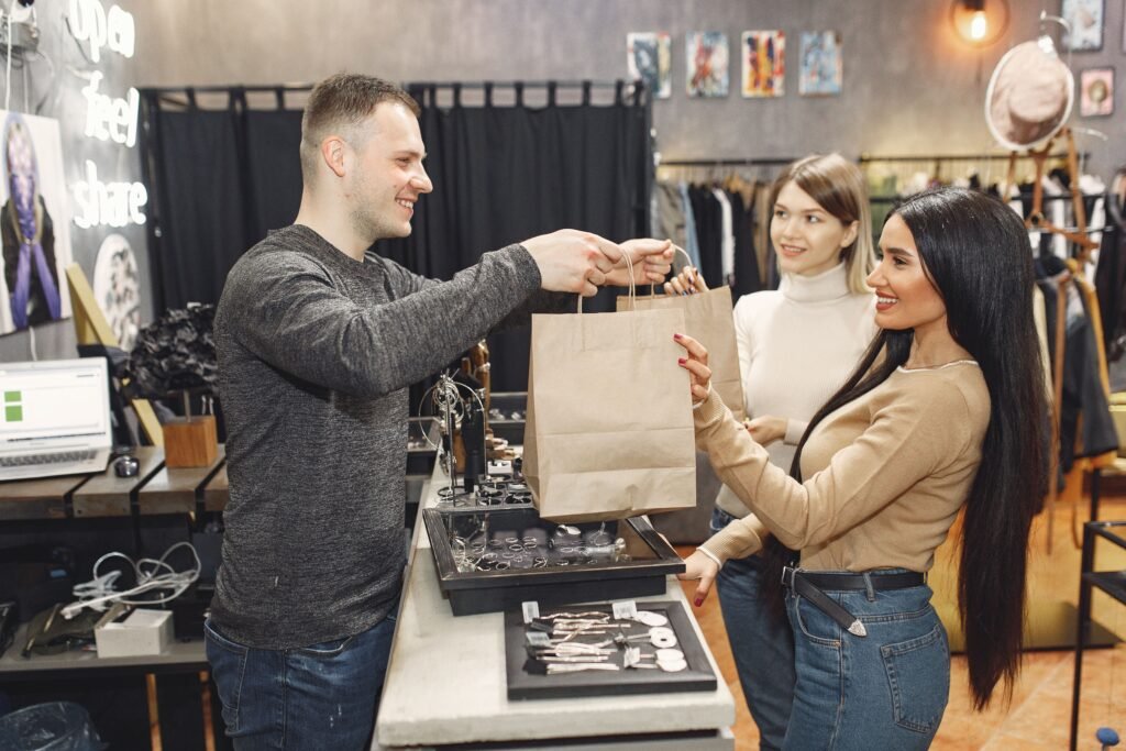 Side view of cheerful male shop assistant standing at counter desk and giving to positive female customers paper bags with purchases in trendy shop